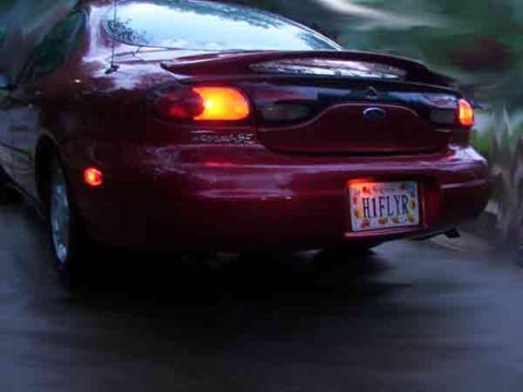 1998 Ford Taurus - Rear view of a red 1998 Ford Taurus sedan driving on a wet road at dusk.