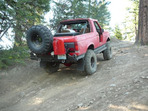 1991 Ford Bronco - Red 1991 Ford Bronco navigates a steep, rocky off-road path with a spare tire and gas can mounted.