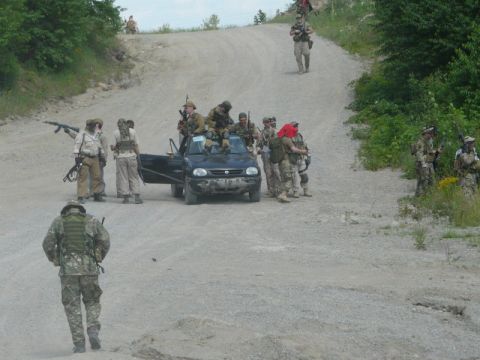 2003 Ford Crown Victoria - A group of people in tactical gear are gathered around a black hatchback on a dirt road, suggesting a staged scene or event.