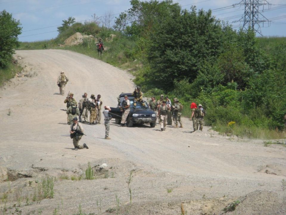 Dark pickup truck on gravel road surrounded by people in camo gear with rifles at an airsoft/milsim event