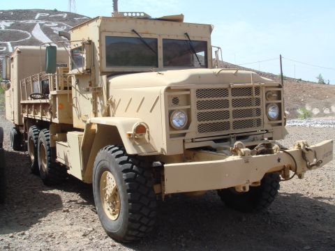 1992 Ford Bronco - A tan military truck is shown from a 3/4 front view, parked on a gravelly surface with a hilly, arid landscape in the background.