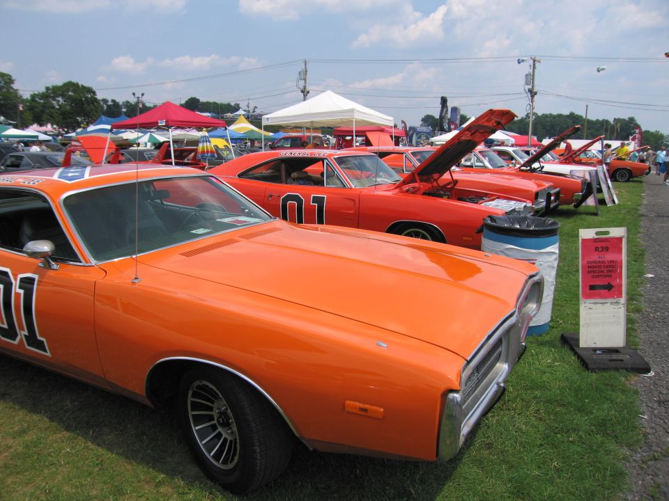 Several orange Dodge Chargers with '01' on doors and Confederate flag on roof parked in a row at an outdoor event