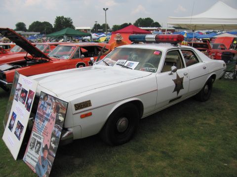 2001 Ford Crown Victoria - A 1977 Plymouth Fury police car is displayed at an outdoor event, with a "Wanted" poster in front of it.