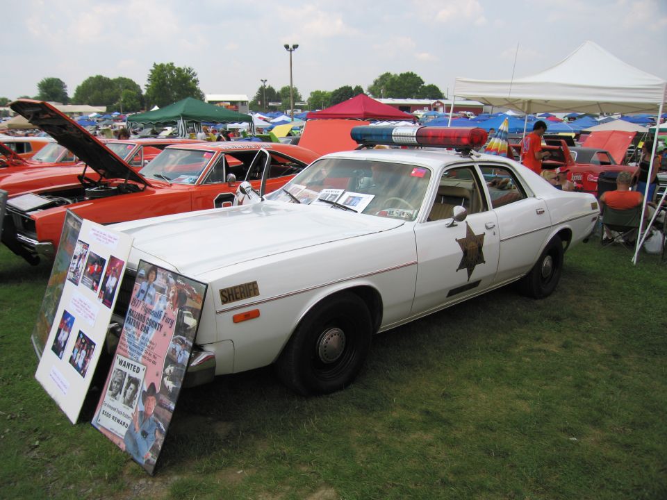 White sheriff car with light bar on top, parked on grass at an outdoor event. Other cars and tents are in the background