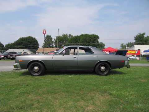 2001 Ford Crown Victoria - A gray 1970s Dodge Monaco sedan is parked on a grassy area with tents and buildings in the background.