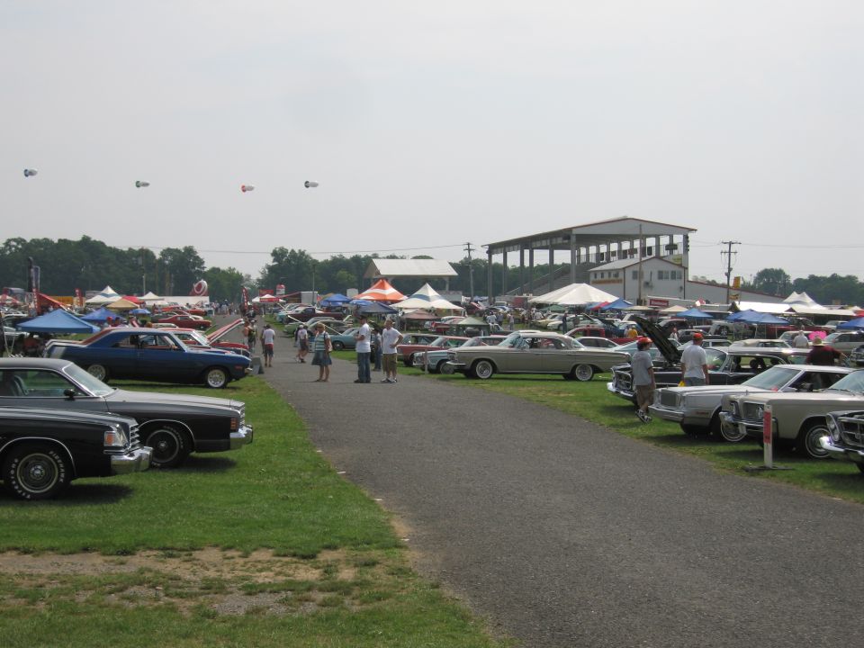 Outdoor car show with many vintage vehicles parked on grass and asphalt, tents set up, and people milling around