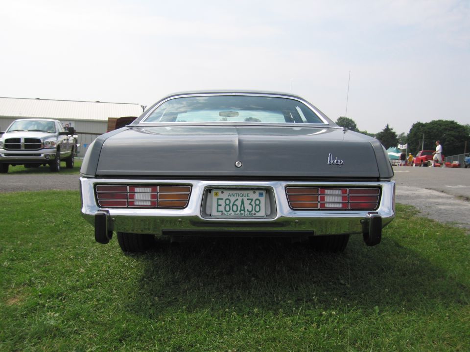 Rear view of a dark gray Dodge with a chrome bumper and a license plate that reads E86A38. A silver pickup truck is parked to the left