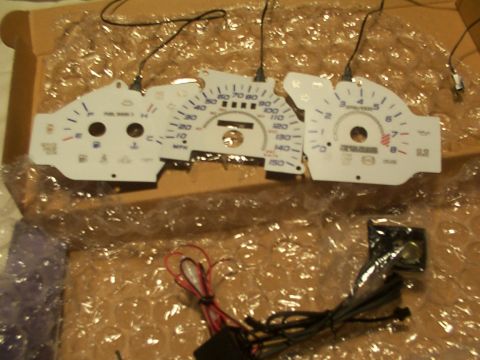 1998 Ford Taurus - A white gauge cluster with black needles and blue markings is shown, with wires attached and resting in bubble wrap.