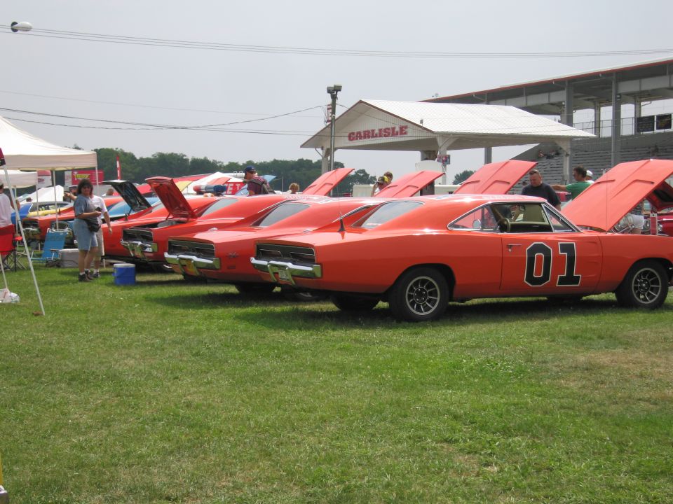 Several orange Dodge Chargers with '01' on the doors parked on grass, hoods up