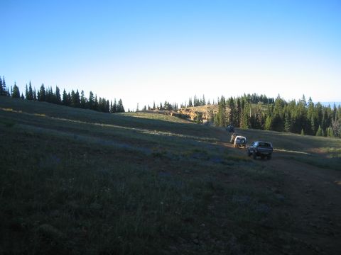 1989 Ford Bronco - A group of off-road vehicles traverse a scenic, sun-drenched hillside under a clear blue sky.
