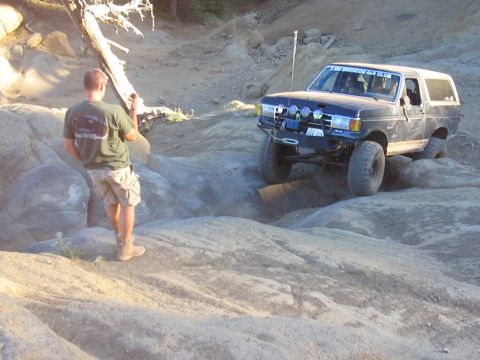 1989 Ford Bronco - A person stands near a white SUV as it drives on a dusty, off-road path.