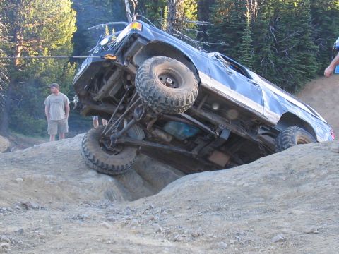 1989 Ford Bronco - A blue 1989 Ford Bronco navigates a challenging off-road obstacle, with one wheel lifted high.