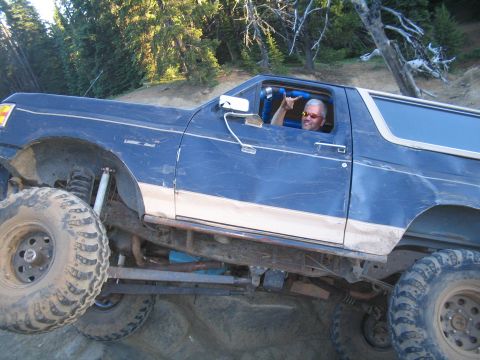 1989 Ford Bronco - A man gestures from the driver's seat of a modified blue SUV on a dirt trail.