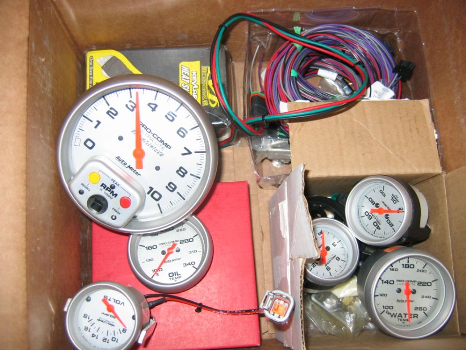 Several automotive gauges, including a tachometer, oil temp, water temp, and voltage meter, are in a cardboard box with wiring