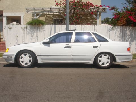 1990 Ford Taurus - This white 1990 Ford Taurus is seen from the side, parked in front of a house with a white picket fence.