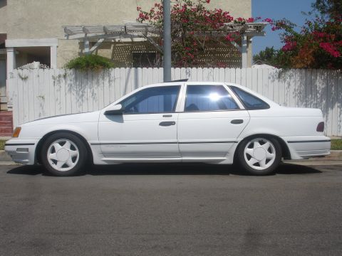 1990 Ford Taurus - A white sedan is parked on the street in front of a white fence and a house.