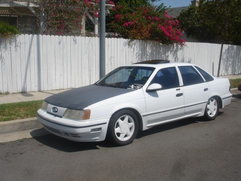 1990 Ford Taurus - A white sedan with a grey hood is parked on the side of the street.