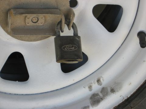 1979 Ford Bronco - A close-up shot shows a dark gray Ford-branded padlock securing a metal bracket attached to a white vehicle wheel. The wheel has triangular…