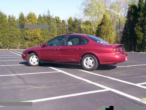 1998 Ford Taurus - This red 1998 Ford Taurus sedan is seen from the side, parked in an empty lot with trees in the background.