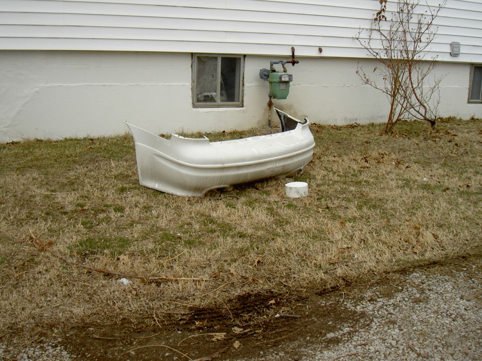 White rear bumper cover removed and sitting on grass beside a house foundation