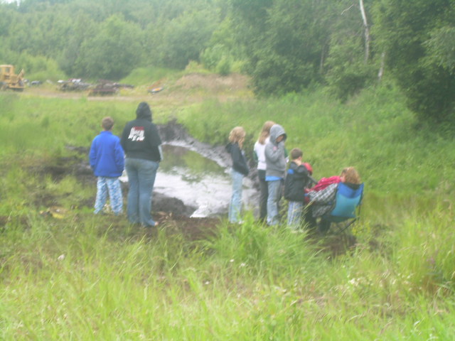 A group of people stand around a muddy area with a bulldozer in the distance.