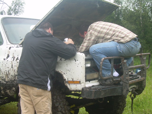 Two men working on the engine of a mud-covered white pickup truck with its hood open.