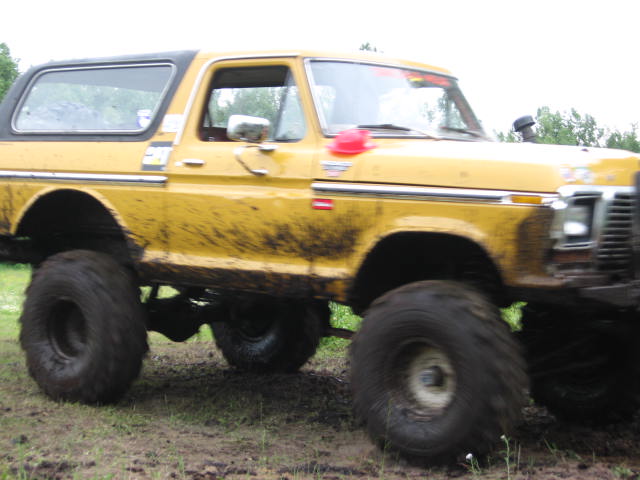 Muddy yellow 1978 Ford Bronco with large off-road tires driving through a field.