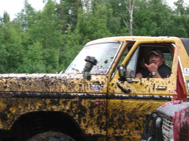 Man in a mud-covered yellow 1978 Ford Bronco, giving a shaka sign.