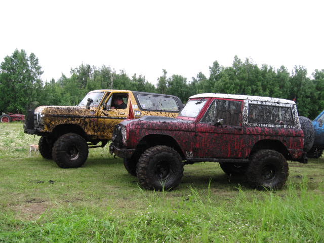 Two mud-covered Ford Broncos, one yellow and one red, parked in a grassy field.
