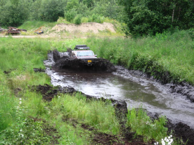 Dark SUV off-roading through a muddy water obstacle, kicking up a large splash.