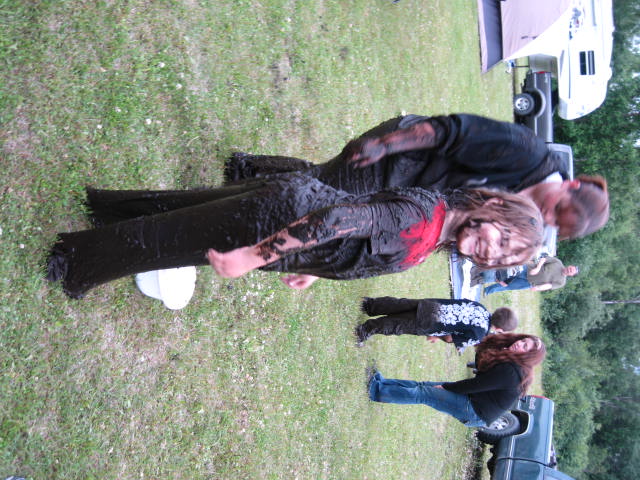 Mud-covered girl smiling, woman next to her, grassy field, bucket.