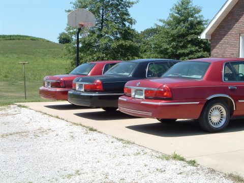 2001 Mercury Grand Marquis - Three Mercury Grand Marquis sedans, two red and one black, are parked in a line on a paved surface. The red car in the foreground shows its…