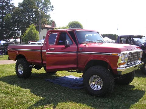 1995 Ford Bronco - A lifted red pickup truck is parked on a grassy area, with another vehicle partially visible to its right.
