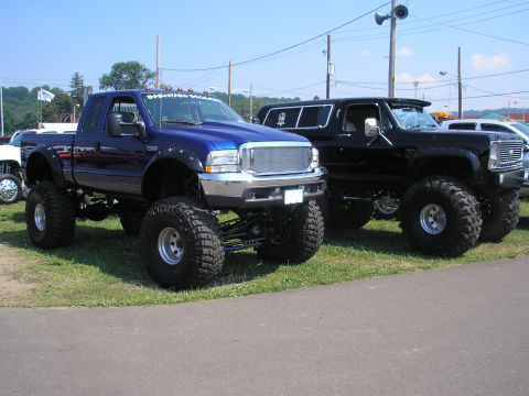 1995 Ford Bronco - Two heavily modified pickup trucks are parked side-by-side on a sunny day.