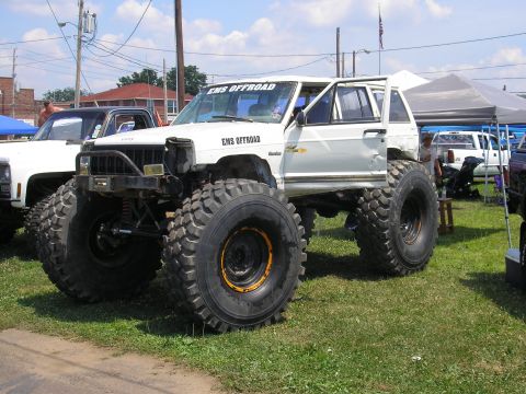 1995 Ford Bronco - A heavily modified white SUV with enormous tires sits on a grassy field next to other vehicles.