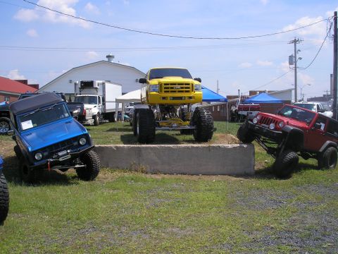 1995 Ford Bronco - Three modified off-road vehicles are parked on grass with buildings in the background.