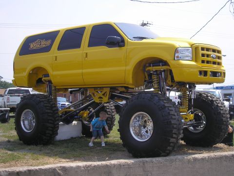 1995 Ford Bronco - This lifted Ford Excursion features massive off-road tires and a custom suspension system, dwarfing a small child nearby.