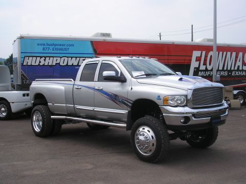 1995 Ford Bronco - A lifted, silver Dodge Ram 3500 dually pickup truck is parked in front of a trailer advertising Hushpower and Flowmaster exhaust systems.