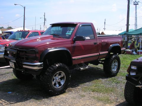 1995 Ford Bronco - A lifted maroon pickup truck is parked on a gravel lot with other vehicles and tents in the background.