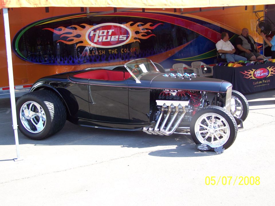 Black custom hot rod with exposed V8 engine and chrome wheels, parked in front of an orange trailer.