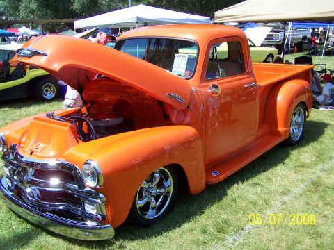 1973 Ford Mustang - A bright orange classic pickup truck is parked on a grassy lawn with its hood open, revealing the engine. The truck has chrome accents and…