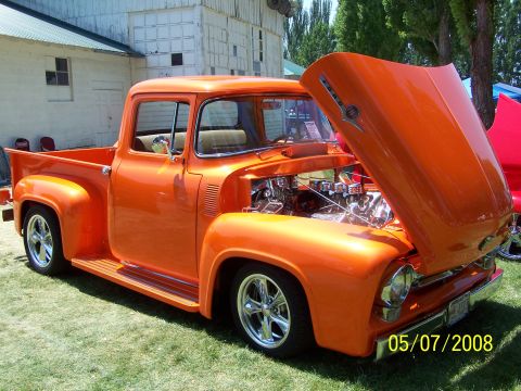 1973 Ford Mustang - A highly customized, bright orange 1950s era pickup truck is parked on a grassy lawn with its hood open, revealing a chrome-laden engine. T…