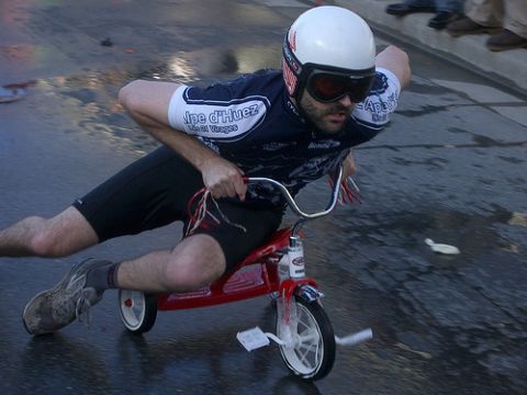 1992 Ford Bronco - A man in a white helmet, red-lens goggles and an 'Alpe d'Huez' cycling jersey rides a child's red tricycle down a wet street, one leg trail…