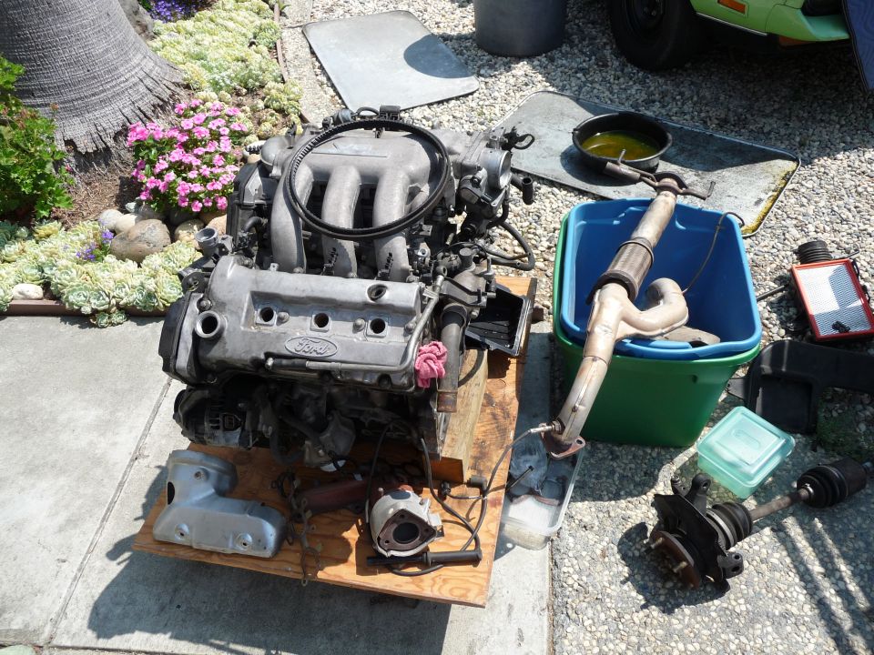Disassembled Ford engine and car parts on a wooden pallet outdoors.