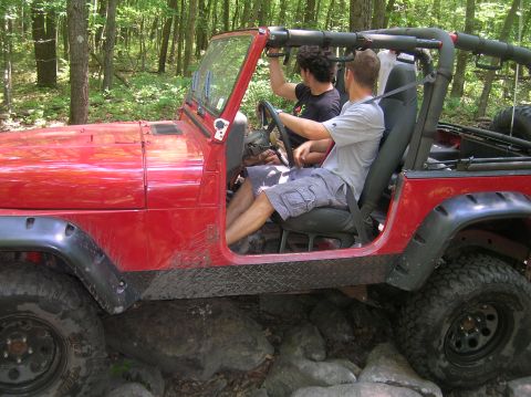 1996 Ford Bronco - Two people navigate a red Jeep Wrangler over a rocky trail in a wooded area.