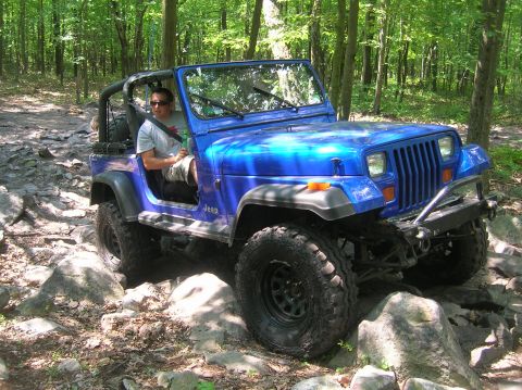 1996 Ford Bronco - A blue Jeep Wrangler navigates a rocky path through a dense forest.