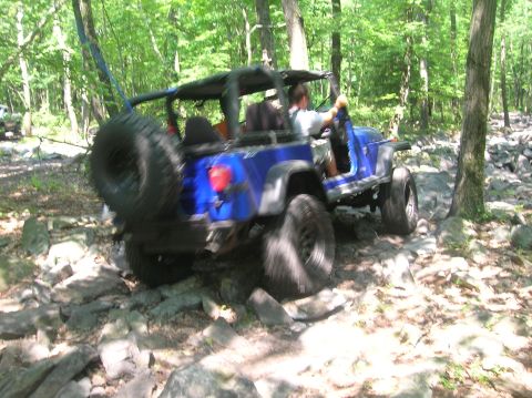 1996 Ford Bronco - A blue off-road vehicle navigates a challenging rocky trail through a wooded area.