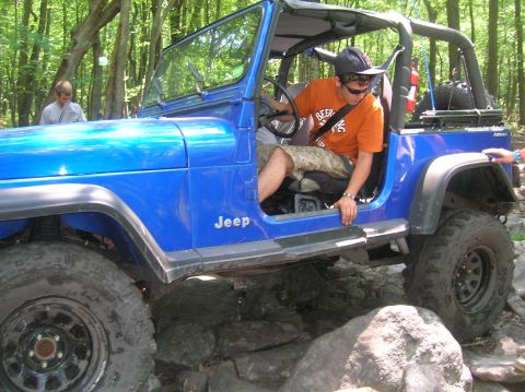 1996 Ford Bronco - A blue Jeep Wrangler is shown off-roading over large rocks in a wooded area.