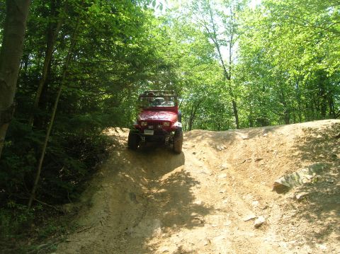 1996 Ford Bronco - A red Jeep Wrangler navigates an off-road trail, climbing a steep dirt incline surrounded by lush green trees.