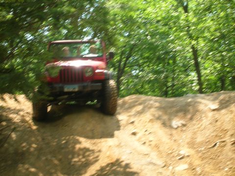 1996 Ford Bronco - A red Jeep Wrangler navigates an off-road trail surrounded by lush green trees.